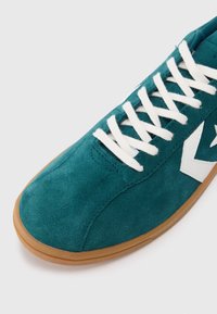 Sapatilha atlética em camurça verde-água com atacadores brancos e um logótipo em forma de chevron branco na lateral, apresentando uma sola de borracha castanha.