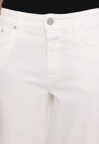 Jean en denim blanc à taille haute, avec une fermeture à bouton argenté, deux poches avant et une étiquette de marque discrète sur la ceinture.