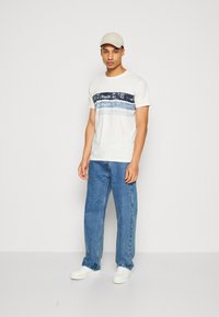 Weißes T-Shirt mit horizontalem blauen Streifenmuster, kombiniert mit locker sitzenden blauen Jeans und weißen Sneakers. Das Modell trägt eine hellbeige Kappe.