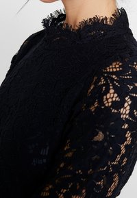 Tummansininen pitsimekko, jossa on korkea kaula-aukko, kukkakuosisia yksityiskohtia sekä läpikuultavat hihat, joissa on monimutkaisia yksityiskohtia ja tekstuuri.