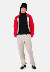 Rote Fleecejacke mit grauen Akzenten, schwarzer gerippter Rollkragenpullover, beigefarbene Cargo-Hosen und schwarze Sportschuhe mit weißen Sohlen.