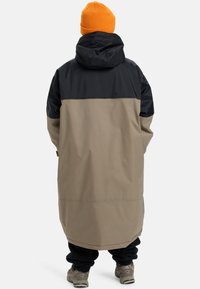 Lange, gewatteerde jas met een zwart bovenstuk en een bruin onderstuk, met een losse pasvorm en waterdicht materiaal, gedragen over donkere broek.