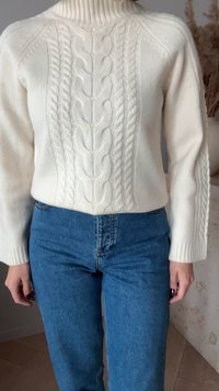 Fehér kötött pulóver, kábel-kötéses dizájnnal, magas gallérral és bordázott ujjakra húzva, testes kék farmerrel párosítva. Puha textúra és laza illeszkedés.
