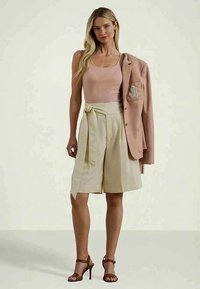 Naine kannab lühikeste varrukatega roosa toppi, beeže vöödiga lühikeseid pükse, pruune kõrge kontsaga sandaale ja roosa bleiserit, mis on tõmmatud ühe õla peale.