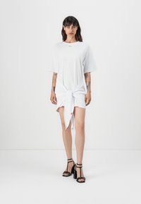 Bílá oversized trikošaty, uvázaná v pase. Obsahuje krátké rukávy, kulatý výstřih a malé logo. Nosené s černými sandály na vysokém podpatku.