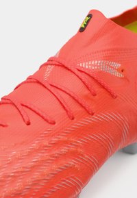 Jasnoczerwony but piłkarski z chropowatą powierzchnią, systemem sznurowania i srebrnymi akcentami po bokach. Wykonany z materiału syntetycznego.