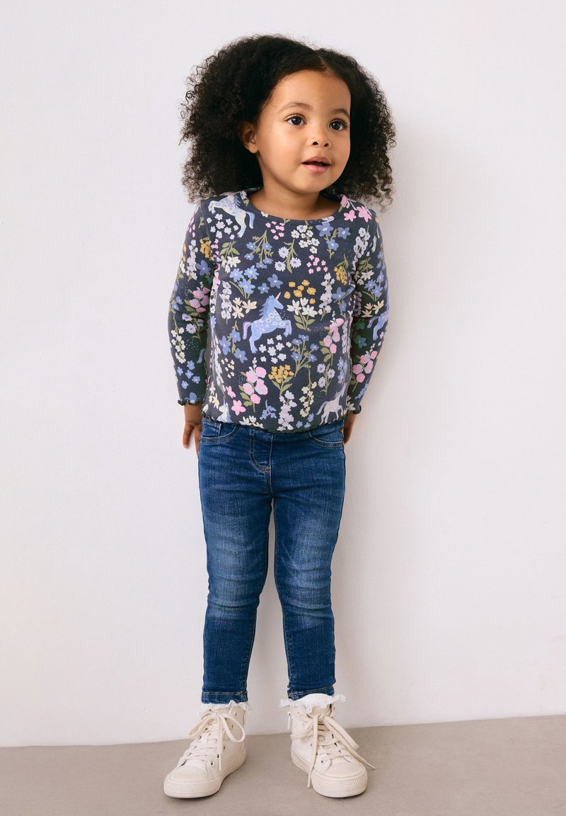 Langærmet grå top med flerfarvet blomstermønster og enhjørning, kombineret med blå jeans og hvide højtop-sneakers med flæsekant.