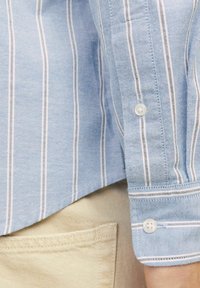 Šviesiai mėlynas marškinėlių apykaklė su baltais ir pilkais vertikaliais dryžiais, turintis mygtuko rankovę ir lygų paviršių, dėvimas ant šviesiai smėlio spalvos audinio.