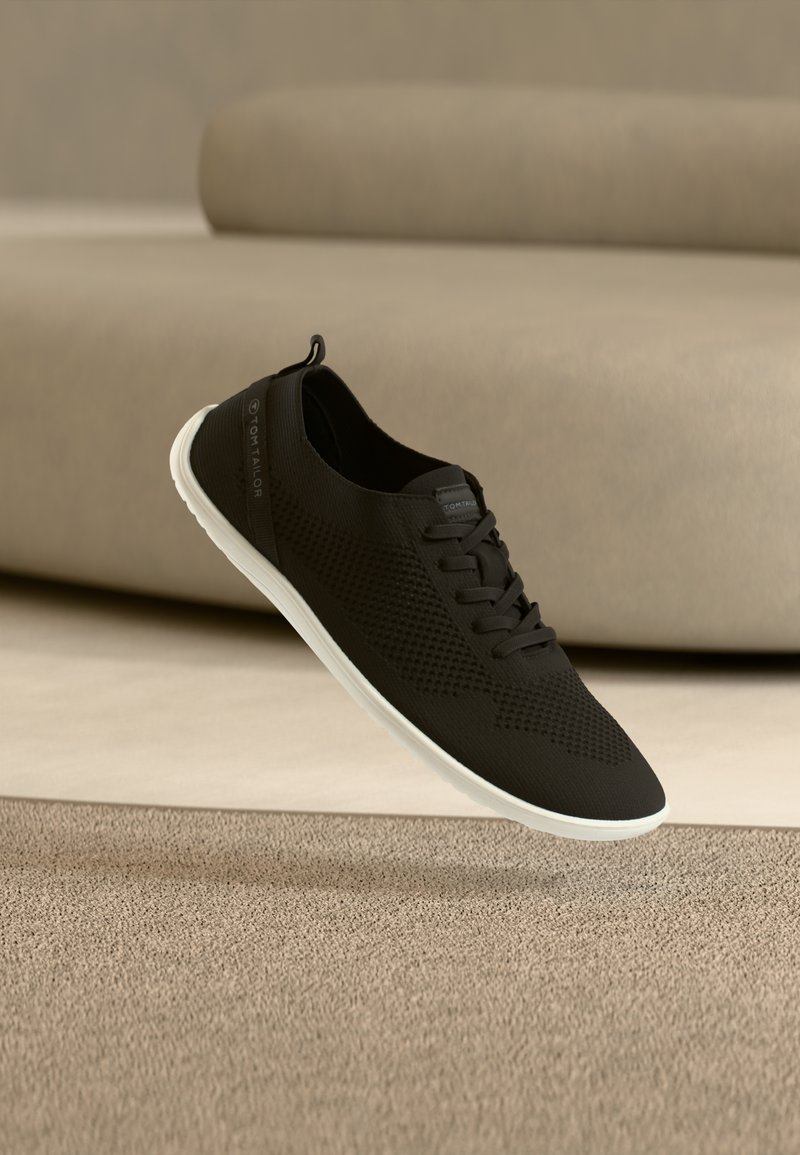 Baskets légères noires à semelle blanche flottant au-dessus d'un tapis beige, avec en arrière-plan un canapé beige flou.