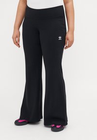 Fekete bővülő sportnadrág ribbed textúrával és széles derékpánttal. Bal csípőn fehér Adidas logó található. Fekete és rózsaszín lábbelivel viselt.