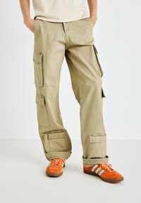 Khakifarbene Cargo-Hose mit mehreren Seitentaschen, geradem Schnitt und weicher Textur, kombiniert mit orangefarbenen Sneakers mit blauen Streifen.