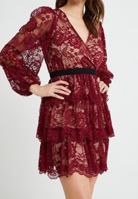 Vestito in pizzo borgogna con maniche lunghe trasparenti, gonna a più strati, scollatura a V e vita nera. Motivo floreale con bordi ondulati.