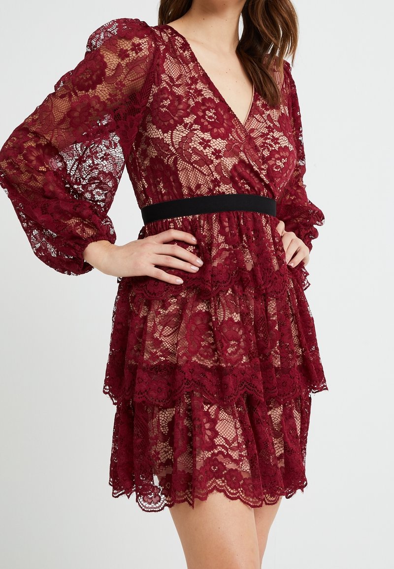 Vestito in pizzo borgogna con maniche lunghe trasparenti, gonna a più strati, scollatura a V e vita nera. Motivo floreale con bordi ondulati.