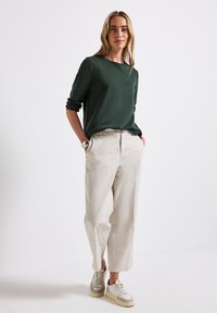 Camicia a maniche lunghe verde scuro, vestibilità comoda; pantaloni larghi color crema; sneakers beige e bianche con accenti testurizzati; sfondo neutro.
