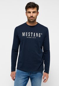 Sötétkék pamut hosszú ujjú póló, elöl fehér "MUSTANG" logóval és a "JEANS COMPANY SINCE 1932" felirattal. Sima tapintású.