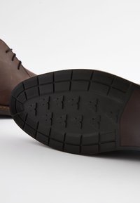 Hnedá kožená topánka s textúrovanou čiernou gumovou podrážkou. Má vzorovaný dezén s trojuholníkovými tvarmi pre lepšiu trakciu.