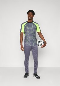 Sivý športový dres s neónovo zelenými akcentmi a vlnkovými vzormi, v kombinácii so sivými nohavicami a čiernymi športovými topánkami, drží futbalovú loptu.