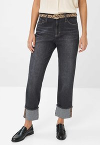 Frau in schwarzen Jeans mit umgeschlagenen Leopardenmuster-Details, einem Leopardenmuster-Gürtel, schwarzen Slippern und einem weißen ärmellosen Oberteil.