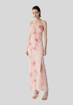 Donna che indossa un abito lungo rosa a fiori con spalline sottili e spacco laterale, abbinato a sandali nude con tacco alto, in piedi davanti a uno sfondo grigio.