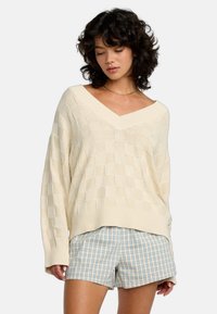 Cremfarbener, strukturiert gestrickter Pullover mit V-Ausschnitt und lockerer Passform, ausgestattet mit einem karierten Muster. Kombiniert mit hellblauen karierten Shorts.