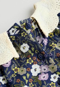 Kvetinová košeľa na gombíky s námorníckym pozadím, krémovým čipkovým golierom a námorníckymi gombíkmi. Obsahuje rôzne malé kvety v purpurovej, žltej a bielej farbe.