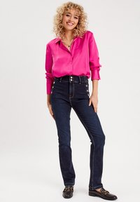Bright pink satin blouse with buttons, paired with high-waisted dark denim jeans featuring gold-tone button accents and a slim fit.