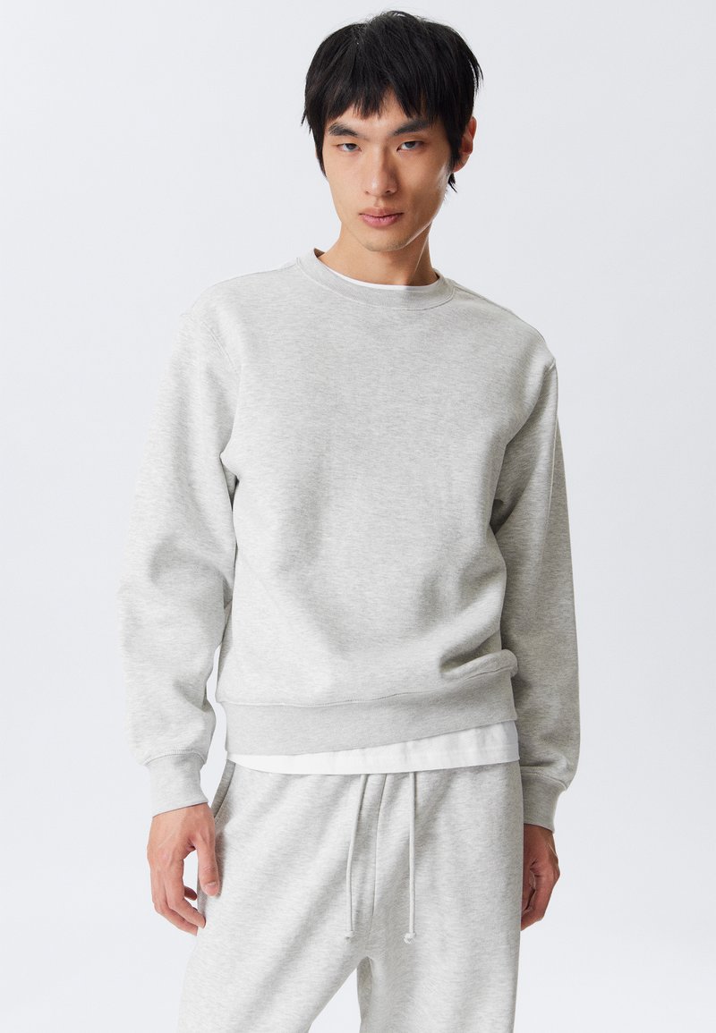 Young man wearing light grey sweatshirt and matching sweatpants stands against white background, looking slightly to the side.