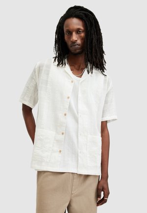 Man met lange dreadlocks die een wit overhemd met korte mouwen draagt over een witte geribbelde tanktop en beige broek voor een effen achtergrond.