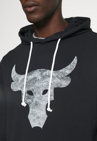 Uomo che indossa una felpa nera con cappuccio e lacci bianchi, con una grande grafica di teschio di toro bianco e usurato sul petto.