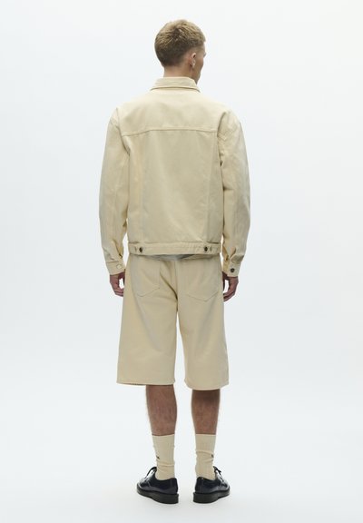 Man wearing beige denim jacket and knee-length shorts, beige socks, and black shoes, standing facing away against white background.