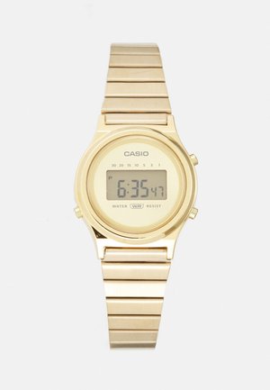 Montre numérique plaquée or avec un cadran rectangulaire, bracelet en segments métalliques liés, et un affichage indiquant l'heure et la résistance à l'eau.