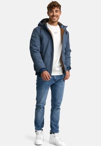 Blå pufferjakke med hætte, brune accenter og lynlås. Hvid sweatshirt under, parret med lyseblå jeans og hvide sneakers.