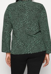 Maglia verde a maniche lunghe con un motivo a foglie nere, vestibilità morbida e scollo rotondo. Il materiale appare liscio con una texture sottile.