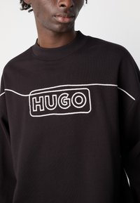 Fekete pulóver kerek nyakkivágással, bordás mandzsettákkal és fehér hímzett "HUGO" logóval elől. Sima textúra, laza szabás.