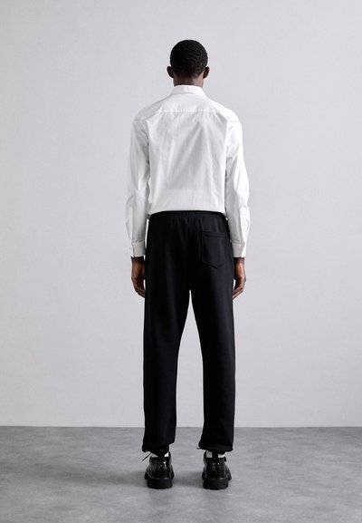Cămașă albă cu mânecă lungă, pantaloni negri cu talie elastică și încălțăminte neagră. Cămașa este netedă; pantalonii au un buzunar lateral și o croială lejeră.