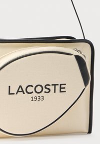 Lacoste taske i cremefarvet tekstureret stof med sorte detaljer. Har en fremtrædende logo og lynlåslukning samt en slank, moderne form.