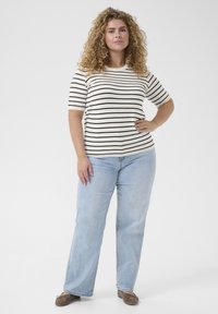Top cu mânecă scurtă în dungi crem cu linii negre, purtat cu blugi evazați de culoare albastră deschisă. Modelul stă cu mâna pe șold, purtând balerini maro.