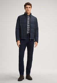 Granatowa pikowana kurtka z wysokim kołnierzem, zamek błyskawiczny i dwie boczne kieszenie, w połączeniu z koszulą w kratę i ciemnymi spodniami. Brązowe botki za kostkę.