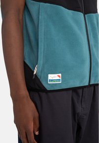 Fleece jacket in teal and black with a zipper, featuring a patch logo and decorative drawcords on pockets. Smooth texture and relaxed fit.