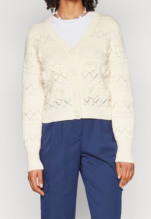 Femme portant un cardigan en maille crème à motifs floraux par-dessus une chemise blanche et un pantalon bleu plissé, debout devant un fond uni.