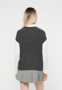 Top grigio lavorato a maglia senza maniche con una texture a coste, indossato sopra una camicia bianca. La parte inferiore presenta un taglio dritto, completato da una gonna plissettata grigia.