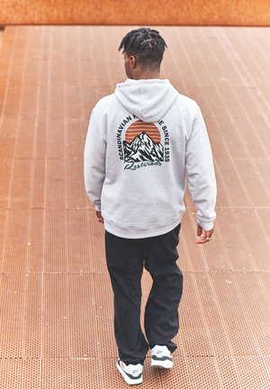 Grå hættetrøje med et bjergdesign og et orange solmotiv på ryggen. Sorte bukser og hvide sneakers med farveaccentuer.