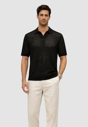 Uomo che indossa una polo lavorata a maglia nera a maniche corte e pantaloni beige chiaro, in piedi con una mano in tasca davanti a uno sfondo semplice.