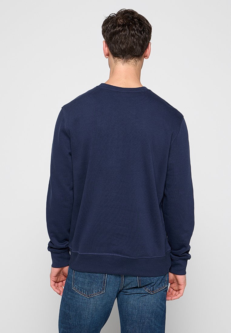 Uomo con capelli corti e scuri che indossa una felpa a maniche lunghe blu navy e jeans blu, in piedi di spalle contro uno sfondo grigio uniforme.