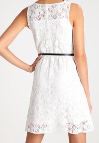 Robe en dentelle blanche avec un corsage ajusté et une fermeture éclair au dos, une ceinture noire à la taille, et une jupe évasée avec un ourlet en forme de vagues.