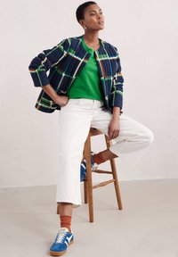 Pikowana granatowa kurtka z kolorowym wzorem w kratkę, zielona bluzka, białe krótki dżinsy, pomarańczowe skarpetki i niebieskie trampki. Drewniane stołek w tle.