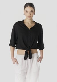 Mladá žena s tmavými vlasmi zviazanými dozadu nosí čiernu košeľu na gombíky zviazanú v páse a biele voľné nohavice, ruky v kapsách, neutrálne pozadie.