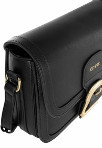 Czarna skórzana torba na ramię o zaokrąglonym kształcie, z złotymi detalami i widocznymi szwami. Posiada klapkę oraz regulowany pasek.