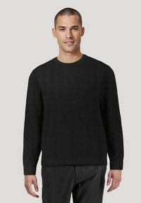 Schwarzer Strickpullover mit einem strukturierten Fischgrätenmuster, rundem Ausschnitt und langen Ärmeln. Getragen mit einer dunkelfarbigen Hose.
