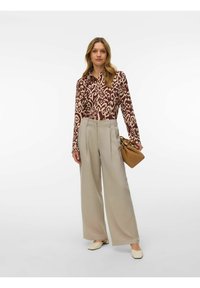 Femme portant une chemise à manches longues à motifs, un pantalon beige à jambes larges, des ballerines crème, et tenant un sac à main marron sur un fond uni.